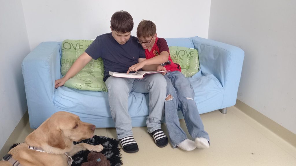 Zwei IntegrationsschĂŒler lesen gemeinsam auf einer Couch dem Schulhund der NĂMS Ebreichsdorf vor