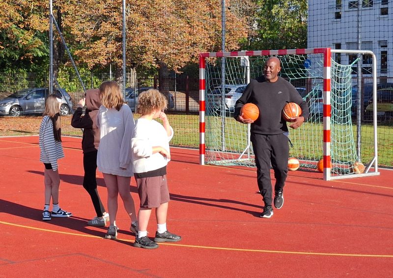Der Coach kommt mit zwei Basketbällen über den Platz auf eine Gruppe Schülerinnen zu