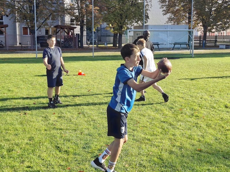 Ein Schüler im blauen Trikot fängt einen Football, hinter ihm läuft ein weiterer Schüler, im Hintergrund lächelt der Coach