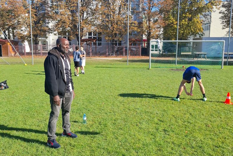 Der Coach steht auf dem Footballfeld, im Hintergrund wärmen sich Schüler auf und üben, umgeben von Herbstbäumen