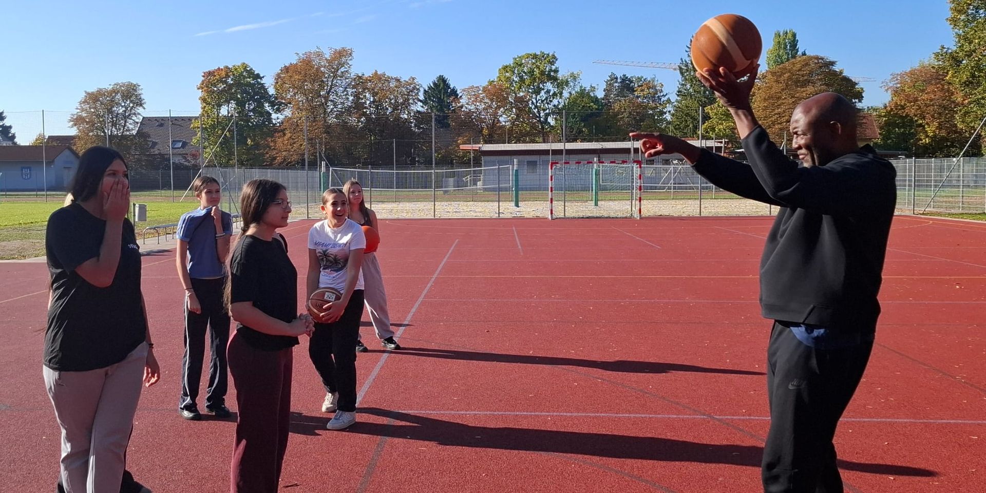 Der Coach hält einen Basketball hoch und erklärt Schülerinnen die Übung auf dem Sportplatz