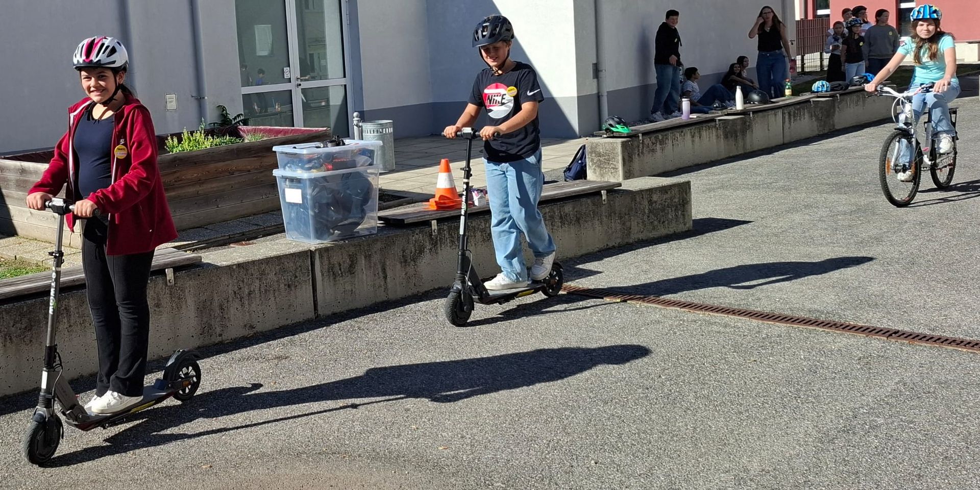 Schülerinnen und Schüler mit Helm auf Scootern und Fahrrad im Parcours auf dem Schulhof