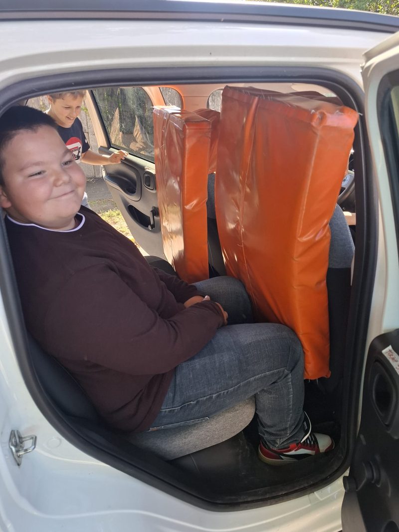 Ein Schüler sitzt lächelnd auf der Rückbank eines Autos, neben ihm stehen orange Polster